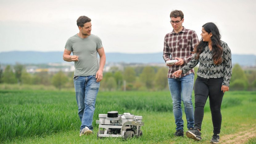 Drei Personen gehen auf einem Feldweg neben einem grünen Feld, eine Person hält ein Tablet, vor ihnen steht ein kleiner Roboter auf dem Weg
