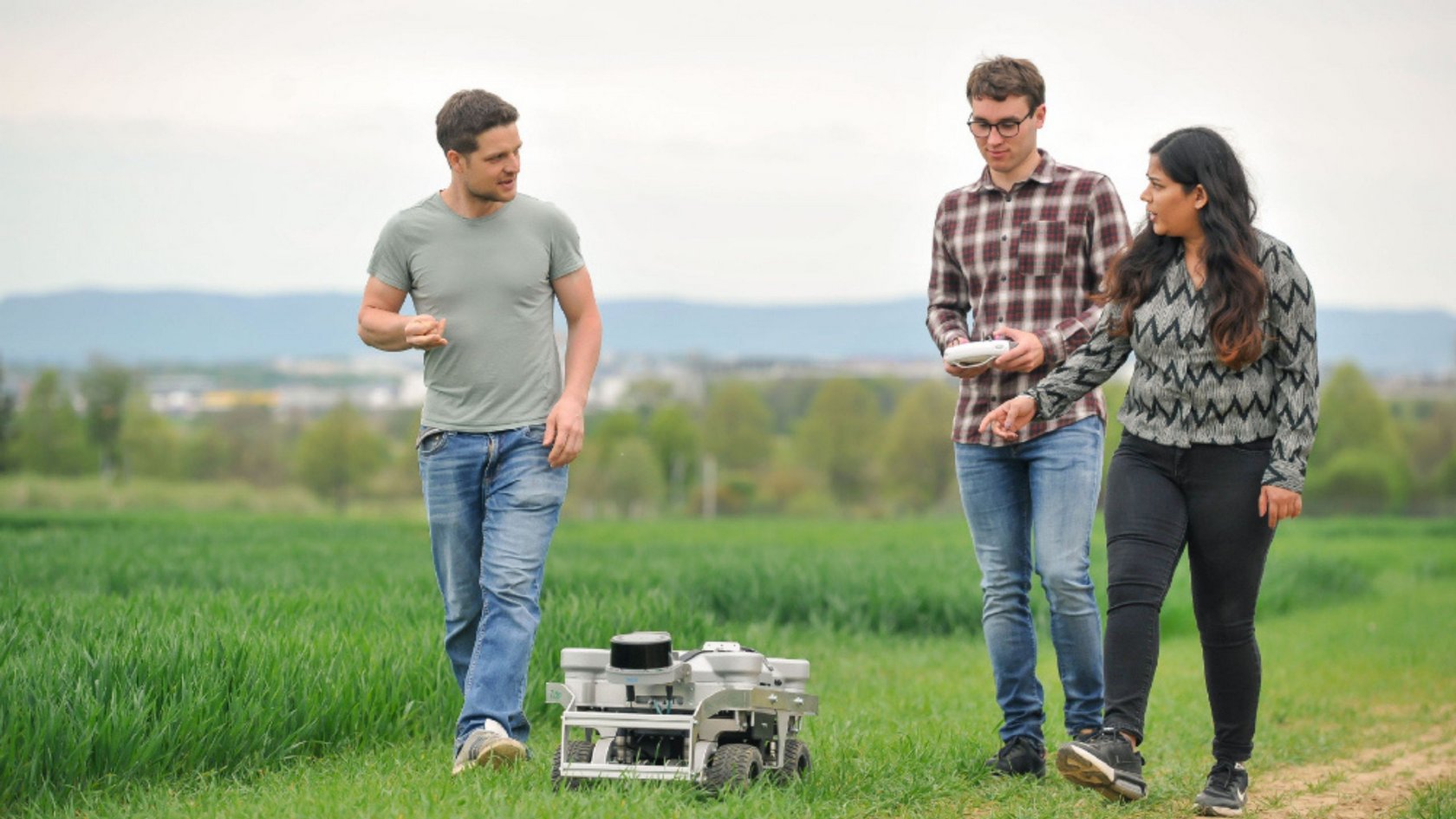 Drei Personen gehen auf einem Feldweg neben einem grünen Feld, eine Person hält ein Tablet, vor ihnen steht ein kleiner Roboter auf dem Weg