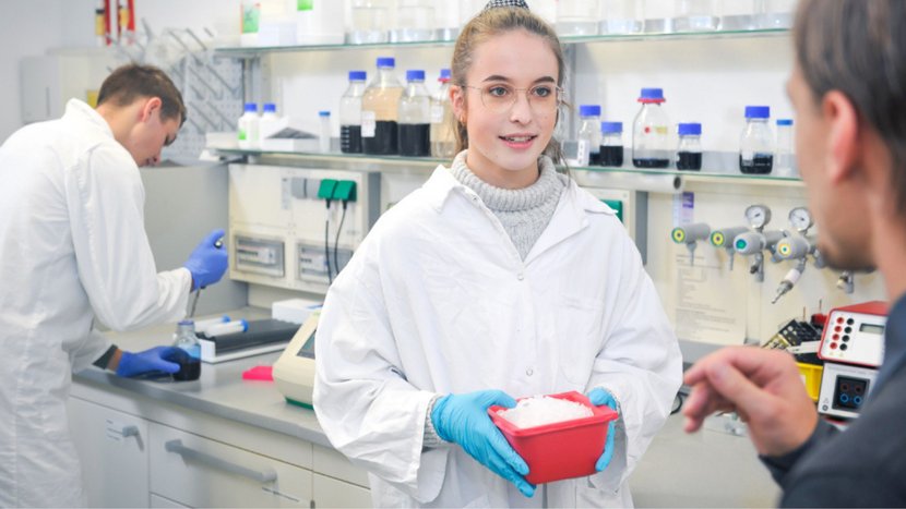 Drei Personen in weißen Laborkitteln und blauen Handschuhen arbeiten in einem Chemielabor, eine Person hält eine rote Kunststoffbox mit weißem Material