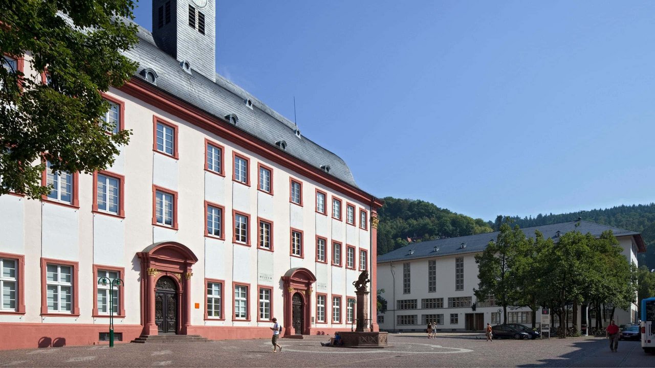 Blick auf zwei historische Universitätsgebäude mit weißen Fassaden, roten Fensterrahmen und Eingängen, umgeben von Bäumen und einem Platz mit Skulptur