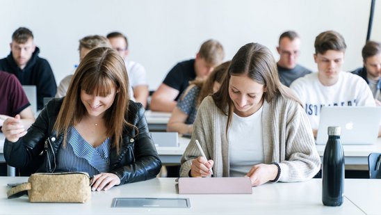 Studierende sitzen in einem Hörsaal an Tischen und schreiben auf Tablets oder Notizblöcken