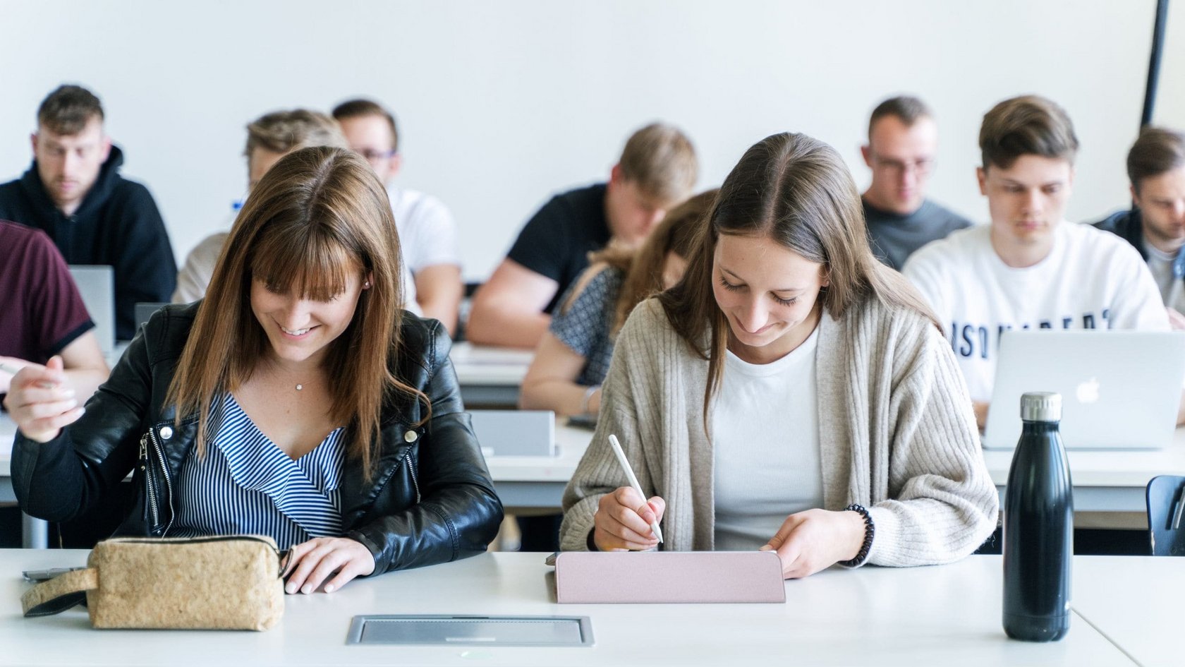 Studierende sitzen in einem Hörsaal an Tischen und schreiben auf Tablets oder Notizblöcken