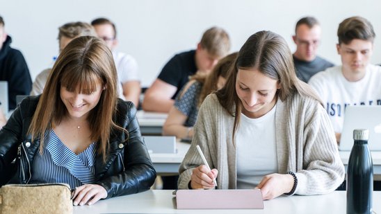 Studierende sitzen in einem Hörsaal an Tischen und schreiben auf Tablets oder Notizblöcken