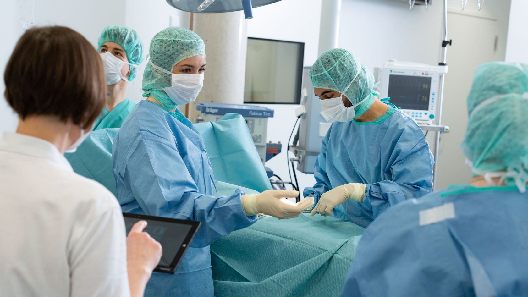 Mehrere Personen in Operationskleidung führen eine medizinische Prozedur in einem OP-Saal durch, eine Person im Vordergrund hält ein Tablet.