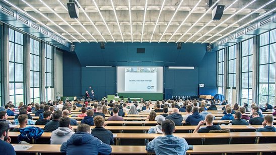 Studierende sitzen in einem Hörsaal in einer Vorlesung, eine Präsentation läuft.