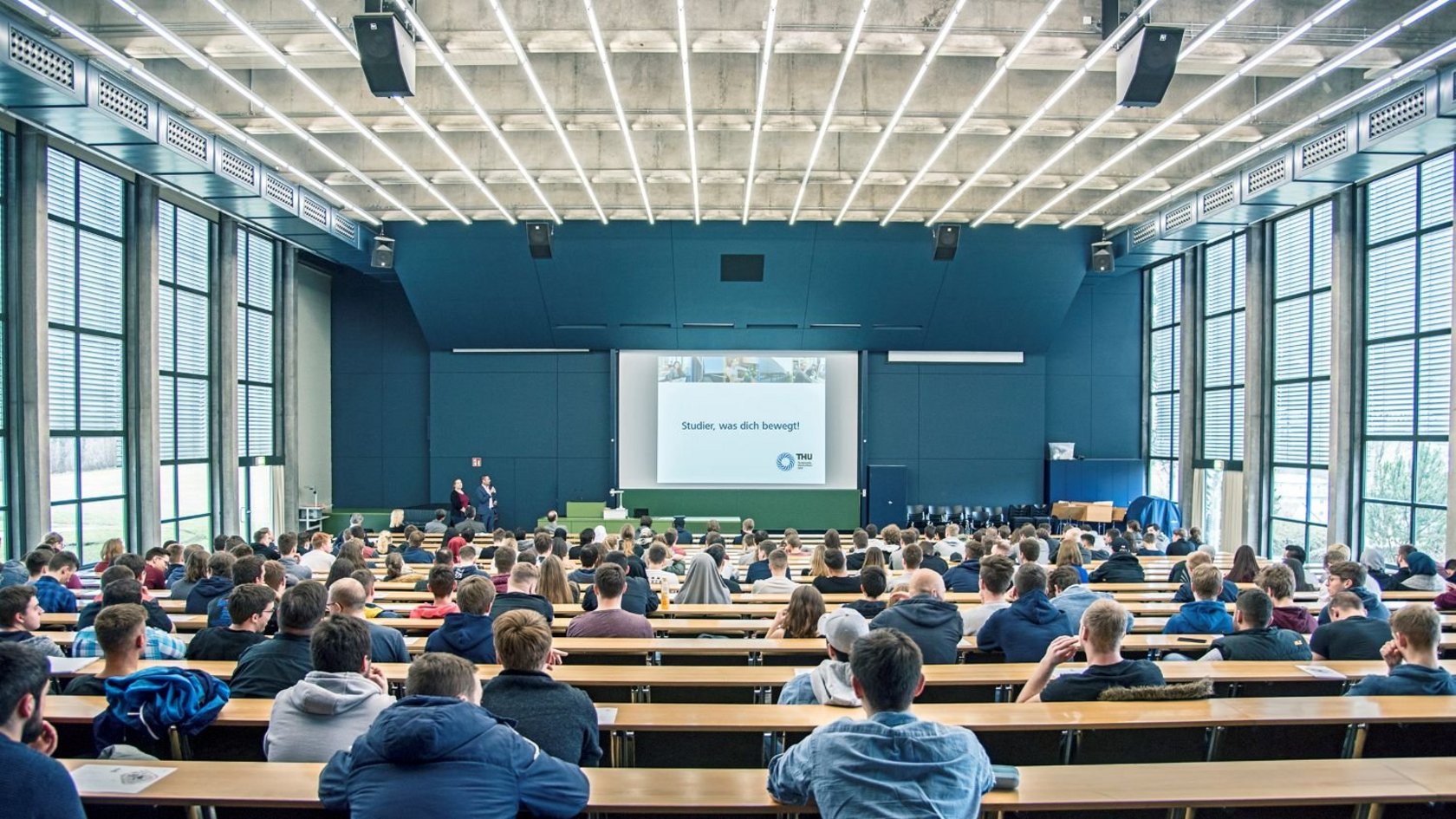 Studierende sitzen in einem Hörsaal in einer Vorlesung, eine Präsentation läuft.