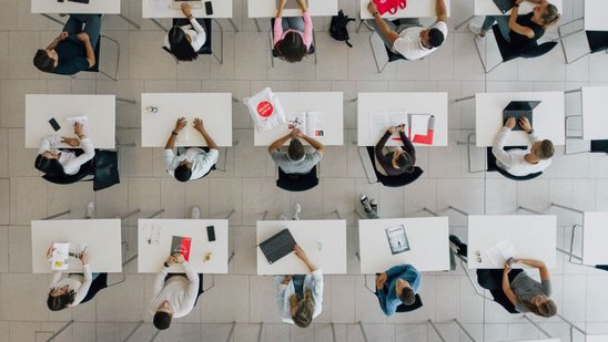 Studierende sitzen in drei Reihen an Tischen mit Notebooks und Arbeitsmaterialien in einem hellen Raum.