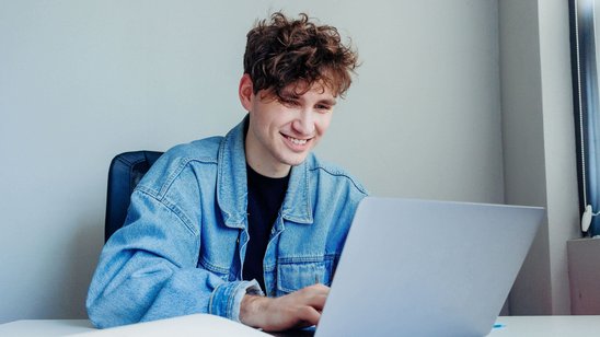 Junger Mann sitzt an einem Tisch und tippt auf einem Laptop, auf dem Tisch liegt ein aufgeschlagenes Buch