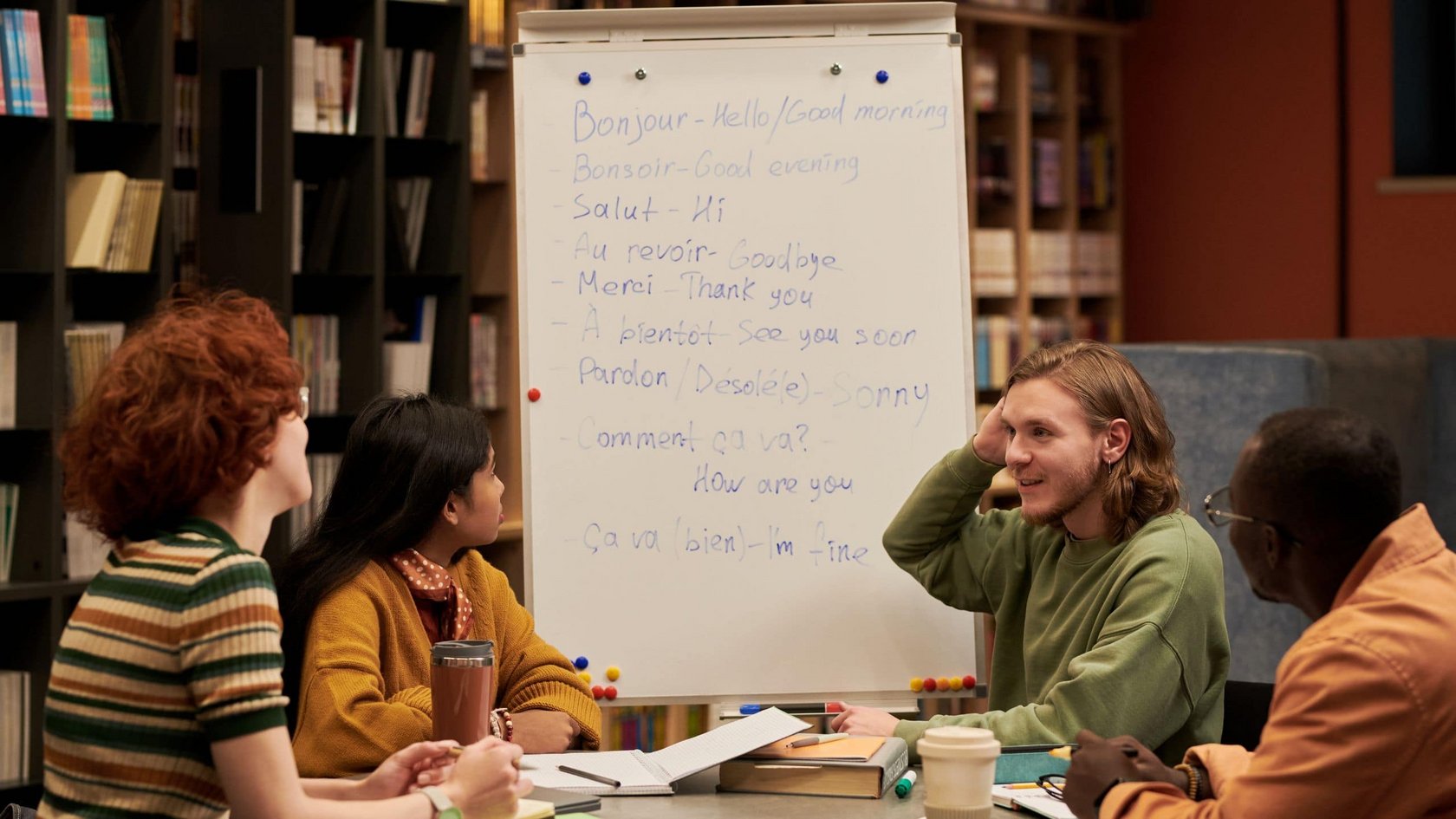 Vier Personen sitzen an einem Tisch vor einem Whiteboard mit französischen Grußformeln und deren englischen Übersetzungen in einem Raum mit Bücherregalen.