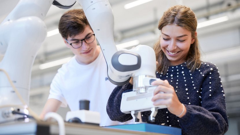 Zwei Studierende bedienen einen Roboterarm in einem Labor oder einer Werkstatt