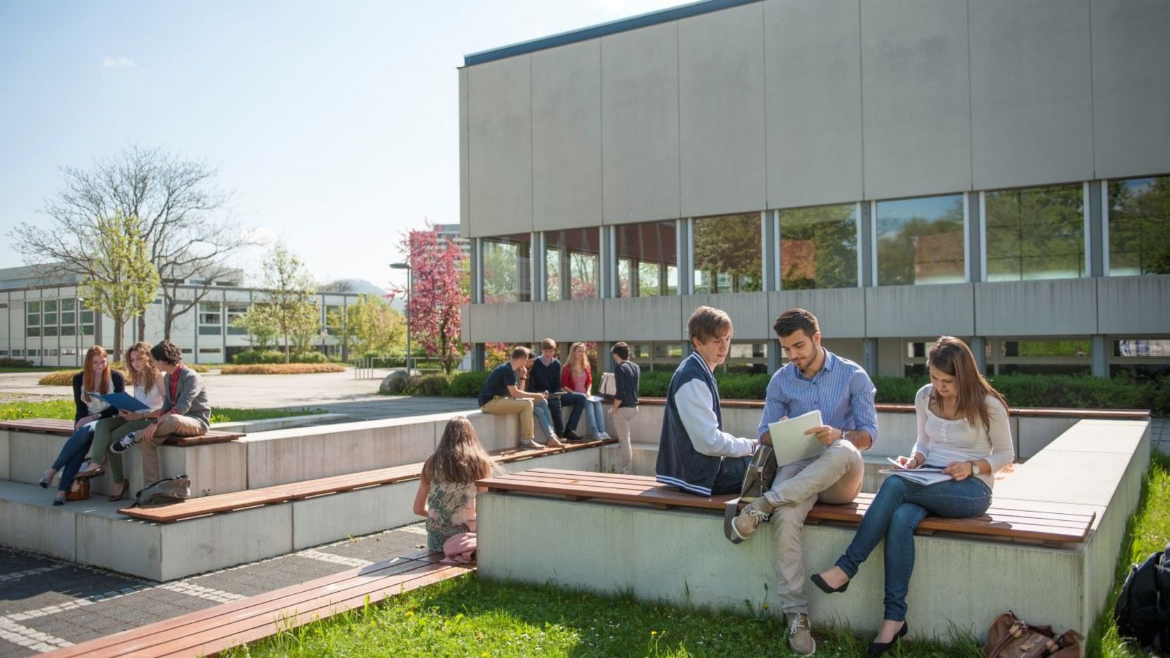 Studierende sitzen auf Bänken vor einen Hochschulegebäude und unterhalten sich.